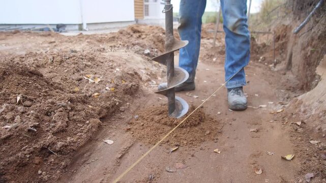 site technician drills soil with auger near trench, technician uses auger for foundation trench earth removal, drilling soil using auger to prepare site foundation and establish trench