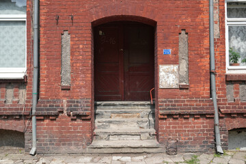 An old, neglected entrance hall of an old stone building with worn steps in the city of Chernyakhovsk, formerly Insterburg. There are two drainpipes and two windows. Background.