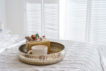 Elegant gift box and ceramic cup on silver tray with Christmas tree lights on cozy white bed in festive decorated bedroom interior. New Year and Christmas atmosphere, festive home decor for holidays
