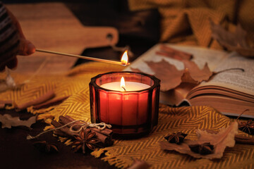 Hand lighting red candle with match surrounded by dried leaves open book cinnamon and yellow fabric