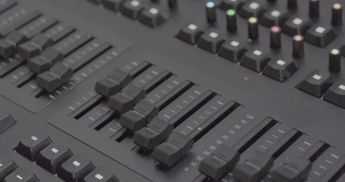 Sound engineer's console, level controls move up and down. Close-up handheld shot, real time speed, artificial indoors lighting.