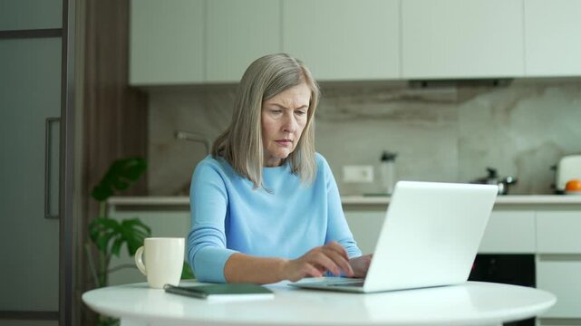 Confused senior female with gray hair having difficulty using laptop sitting at the kitchen table at home. Worried puzzled elderly woman has a problem complexity working on application on computer