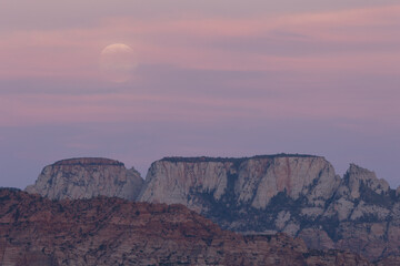 Pink and purple clouds almost hide the full moon rising above the western face of the Altar of Sacrifice peak in Zion NP Utah USA on a September evening just after sunset.
