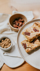 A plate of two brownies with pecans on top sits on a table next to a bowl of pec