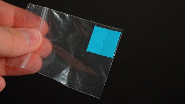 Hands of the drug dealer show off LSD strip blotter in small plastic bag. Dry drops of Lysergic acid drug on paper tabs lucy for sale in club, party and street. Illegal drugs trafficking.