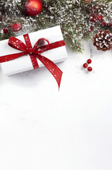 Christmas gift with red ribbon, balls and spruce branches on snow covered surface, snowfall and white wooden board abstract background. Top view, copyspace.