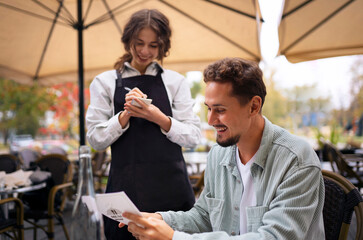 Man reading menu at outdoor cafe with waitress standing nearby ready to take order at the restaurant