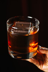 Dark and elegant cocktail in a glass with ice and a piece of chocolate, held by a hand on a dark red background. Sophisticated drink concept, stylish bar or lounge atmosphere.