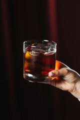 Dark and elegant cocktail in a glass with ice and a piece of chocolate, held by a hand on a dark red background. Sophisticated drink concept, stylish bar or lounge atmosphere.