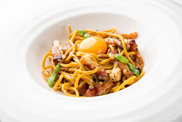 Spaghetti carbonara in a white bowl with crispy pork and a raw egg yolk on top, garnished with...