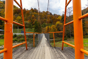 Suspension bridge with orange rails in autumn forest. A narrow wooden suspension bridge with bright orange railings stretches across a river into a vibrant autumn forest with colorful trees.