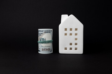 White ceramic house model beside rolled hundred dollar bills on black background, symbolizing investment, security, and financial stability. copy space
