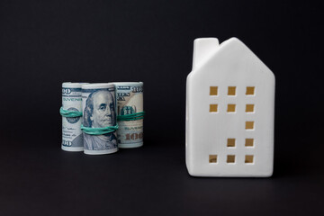 Stacked rolls of cash beside a white ceramic house model on a dark background, symbolizing finance and real estate investment opportunities. copy space