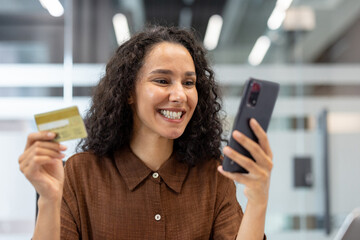 Happy young woman smiling while holding smartphone and credit card, making a secure mobile payment or shopping online with joy and confidence in convenient digital commerce