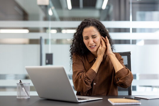 Young woman at her office desk clutching her ear in pain, wincing from earache or tinnitus while working on a laptop, stressed and uncomfortable at work