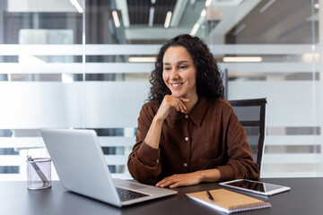 Young woman with curly hair sitting at an office desk, happily engaging in a virtual meeting or online conference on her laptop, demonstrating remote work and communication