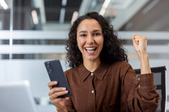 Happy young businesswoman raising her fist in triumph after receiving good news or achieving a goal, celebrating a victory while holding a smartphone in a modern office setup