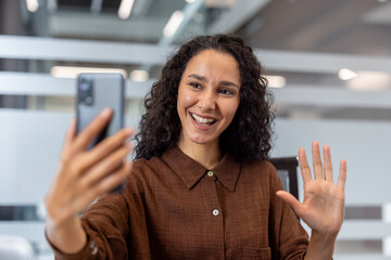 Young smiling woman with curly hair is waving hello while engaging in an online video conference call using her mobile phone, in a modern office environment