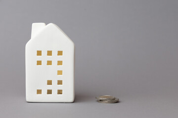White ceramic house model beside coins on gray background, symbolizing finance, investment, and the concept of home ownership in modern economy. copy space