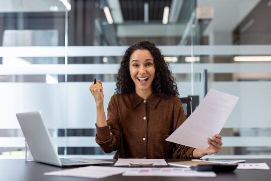 Young mixed race businesswoman celebrating achievement and feeling excited, holding success document and gesturing with a joyful expression while working in a bright office environment