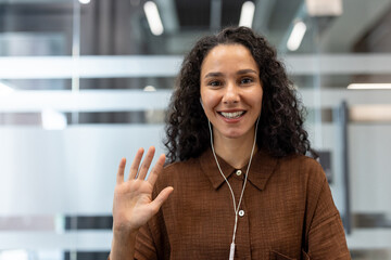 Smiling young woman with curly hair wearing wired earphones, waving hand and looking at camera during an online video call from a modern office environment