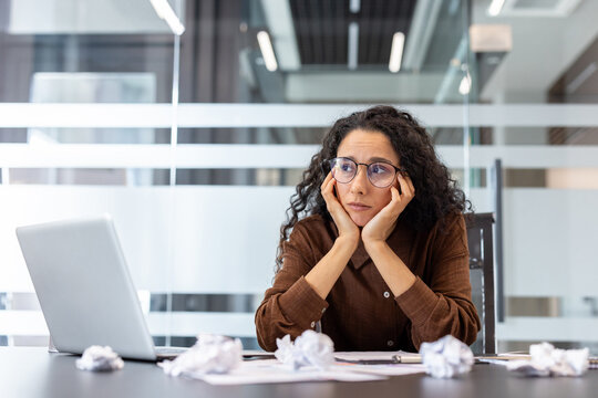 Woman feeling overwhelmed at her office desk, looking frustrated and tired, surrounded by crumpled papers, dealing with work pressure, deadlines, and a lack of inspiration - Powered by Adobe