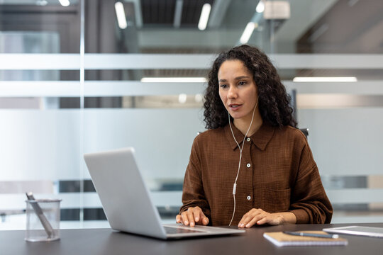 Professional woman focused on a laptop during an online video conference in a modern office, wearing wired earphones while participating in a virtual meeting and working remotely