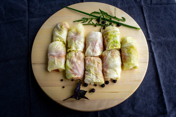Stuffed cabbage leaves stuffed with meat and rice, semi-finished product, lies on a wooden round board, decorated with dill, onion, garlic, basil, arugula, peppercorns