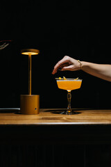 Stylish yellow cocktail in an elegant glass on a wooden bar with warm lighting and a golden lamp. Minimalist scene with a woman's hand reaching for the drink.

