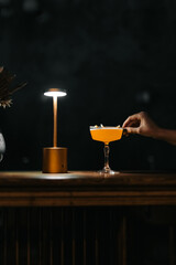 Stylish yellow cocktail in an elegant glass on a wooden bar with warm lighting and a golden lamp. Minimalist scene with a woman's hand reaching for the drink.
