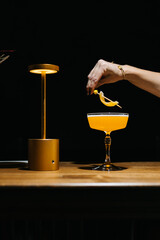 Stylish yellow cocktail in an elegant glass on a wooden bar with warm lighting and a golden lamp. Minimalist scene with a woman's hand reaching for the drink.
