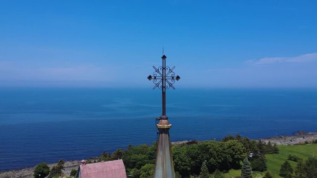 A wrought iron cross sits atop a church steeple, facing a merging ocean and blue sky on the horizon. Sainte-F&eacute;licit&eacute; Church, Quebec, Canada, 2025.