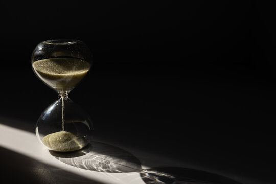 Hourglass with golden sand on dark background illuminated by a sunbeam. Aging concept. Selective focus and copy space