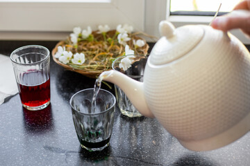 A hand pours hot water from a white teapot into a glass with dye, preparing to dye Easter eggs