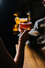 Two people holding colorful cocktails in elegant glasses, one yellow with garnish and one pink with berry, in a cozy bar atmosphere. Celebration and lifestyle concept.
