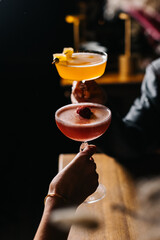 Two people holding colorful cocktails in elegant glasses, one yellow with garnish and one pink with berry, in a cozy bar atmosphere. Celebration and lifestyle concept.
