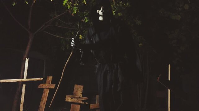 A classic grim reaper in a hood with skeleton head and sickle lurking in a cemetery at night and approaching the viewer. Vintage horror or Halloween death in a graveyard.