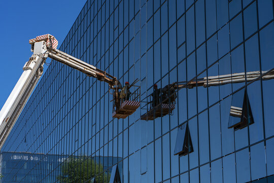 A construction crane with a cradle or lifting platform is reflected in the fa&ccedil;ade glazing of an office building. A construction worker in the crane bucket works on the fa&ccedil;ade glazing.