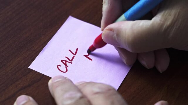 A hand writes text Call Me on a small purple note with a red marker