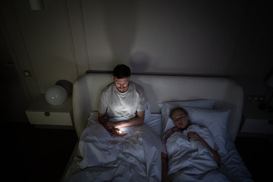 Tired man using smartphone lying in bed at night while wife sleeps beside him in dark bedroom high angle. Compulsive phone use at night, modern insomnia, screen time problem, solitary wakefulness