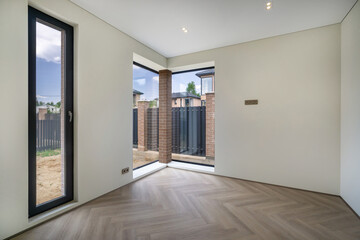 A bright room showcases large windows and an open layout in a newly built home. Light floods in, highlighting the elegant flooring and contemporary walls, creating a welcoming atmosphere.