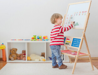 Cute little child playing at a magnetic board, learning letters, numbers and geometric shapes at home. Educative and funny indoor activity for early development.