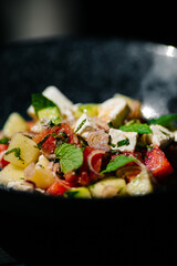 Fresh summer salad with tomato, cucumber, feta cheese, red onion and mint leaves served in a black bowl. Healthy vegetarian meal, gourmet restaurant food concept.
