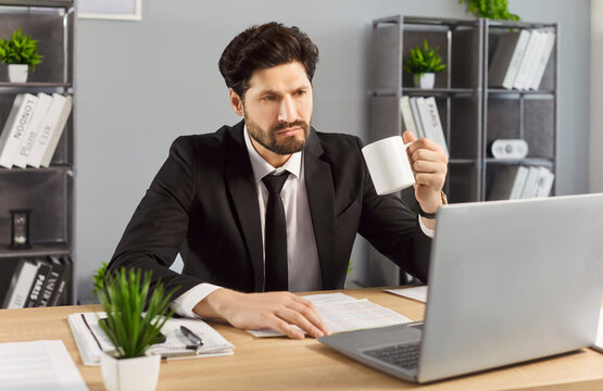 Business man in office with coffee looking at laptop. Wearing suit, professional employee manager checks paperwork while working at the desk. Clear concept of corporate focus and productivity.