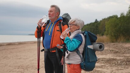 Hiking tourism adventure. Senior couple man woman drinking water enjoying outdoor recreation hiking on beach. Old people backpackers hikers enjoy walking hike trekking active vacation beauty of nature - Powered by Adobe