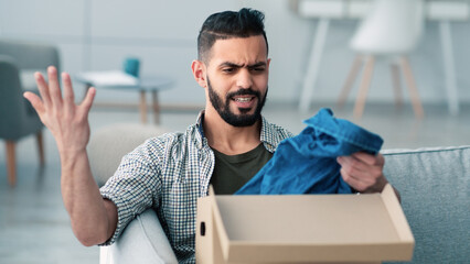 A man sits on a sofa in a stylish living room, looking upset as he examines a piece of clothing just taken out of a delivery box. His expression shows disappointment or confusion.