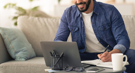 A man sits comfortably on a sofa in a well-lit living room, focused on his laptop while jotting notes in a notebook. A cup of coffee and a phone are on the table beside him.