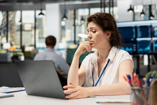 White female manager checking analytics on device, analyzing financial data with puzzled expression about company information. Concerned woman with hand on chin reviewing documents on laptop in office