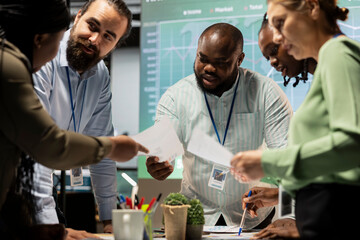 Night time meeting unfolds as a multiethnic team gathers in a strategy room, analyzing visual data, statistics and risk assessment files to drive project development forward for efficiency.
