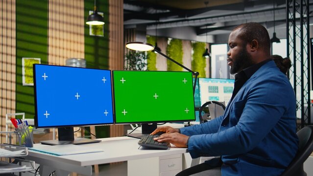 African american businessman examines reports with isolated dual monitors, building a new business strategy for global development. Objectives alignment with productivity.